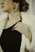 A person wearing elegant jewelry including gold earrings, rings, a bracelet, and a necklace. The individual is dressed in a black ribbed top and a white fur coat, with their hair styled in a bun.