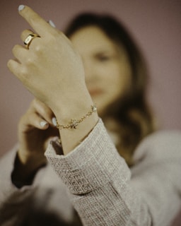 A person wearing a light-colored textured sweater is focusing on displaying a wrist adorned with a delicate gold bracelet featuring small charms. The hand is raised, emphasizing the jewelry, with a ring also visible on one finger. The background is softly blurred, highlighting the bracelet.
