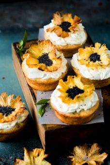 Cupcakes topped with creamy white frosting and decorated with dried pineapple flowers and black seeds. The cupcakes are arranged on a wooden tray with a few mint leaves for garnish.