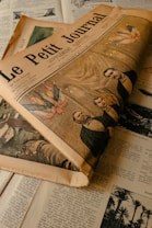 A vintage newspaper titled 'Le Petit Journal' is placed on top of various pages filled with text and illustrations. The main newspaper displays an illustration on the cover, featuring three men and some flags. The overall color scheme is muted and aged, enhancing the historic feel of the image.