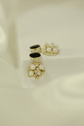 Elegant jewelry pieces featuring intricate designs with pearls and gold accents are displayed against a soft, textured background. The jewelry includes earrings that combine black stones and clusters of pearls with shiny embellishments.