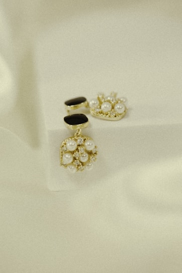 Elegant jewelry pieces featuring intricate designs with pearls and gold accents are displayed against a soft, textured background. The jewelry includes earrings that combine black stones and clusters of pearls with shiny embellishments.