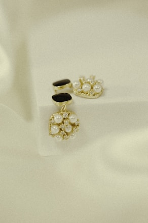 Elegant jewelry pieces featuring intricate designs with pearls and gold accents are displayed against a soft, textured background. The jewelry includes earrings that combine black stones and clusters of pearls with shiny embellishments.