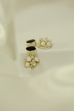 Elegant jewelry pieces featuring intricate designs with pearls and gold accents are displayed against a soft, textured background. The jewelry includes earrings that combine black stones and clusters of pearls with shiny embellishments.