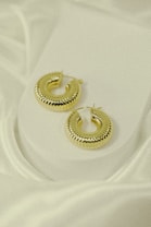 A pair of gold hoop earrings with a ridged texture is placed on a white fabric background. The earrings have a glossy, reflective surface that catches the light, highlighting their intricate design. The soft folds of the fabric add an elegant and luxurious feel to the composition.