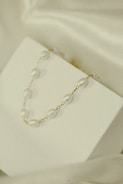 A delicate necklace with pearl-like beads and gold links is elegantly displayed on a minimalistic white surface. The soft lighting and subtle shadows add to the understated elegance.