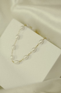 A delicate necklace with pearl-like beads and gold links is elegantly displayed on a minimalistic white surface. The soft lighting and subtle shadows add to the understated elegance.