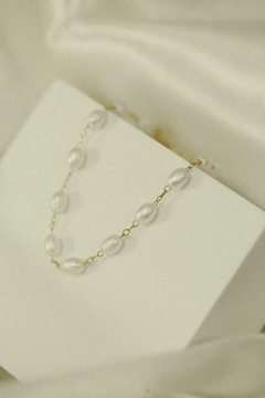 A delicate necklace with pearl-like beads and gold links is elegantly displayed on a minimalistic white surface. The soft lighting and subtle shadows add to the understated elegance.