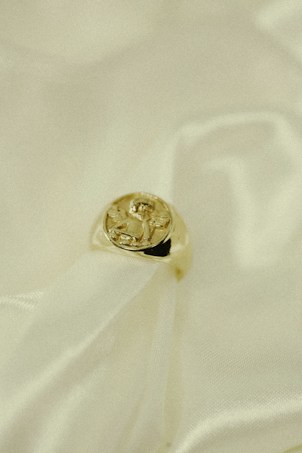A gold ring with an intricate design on its top rests gently on a soft, creamy fabric. The fabric has a flowing texture, emphasizing the luxurious feel of the composition.