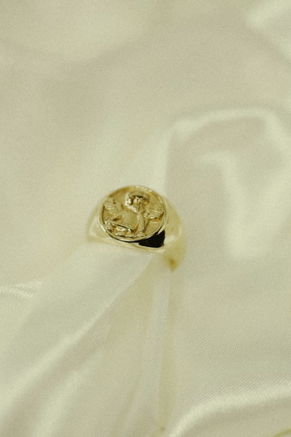 A gold ring with an intricate design on its top rests gently on a soft, creamy fabric. The fabric has a flowing texture, emphasizing the luxurious feel of the composition.