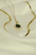 A gold necklace with an ornate pendant featuring a dark gemstone is laid out on a soft, light-colored fabric. The pendant is surrounded by a series of small, sparkling stones set in a circular pattern. The necklace chain is elegantly curved and rests softly against the fabric.