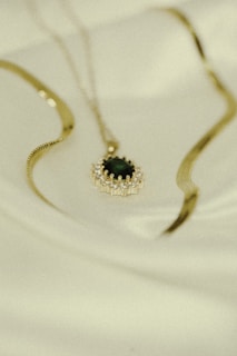 A gold necklace with an ornate pendant featuring a dark gemstone is laid out on a soft, light-colored fabric. The pendant is surrounded by a series of small, sparkling stones set in a circular pattern. The necklace chain is elegantly curved and rests softly against the fabric.
