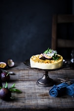 A cheesecake garnished with fresh figs and mint leaves is presented on a dark metal cake stand. The setup is placed on a rustic wooden table. In the background and foreground are whole and halved passion fruits, while a rolled-up blue napkin lies beside them.