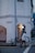 An elderly man pushes a shopping cart along the sidewalk in front of a white colonial-style building with arched doorways. A bicycle is parked nearby, and another man is standing beside a green trash bin. The scene is set during twilight, giving it a calm and serene atmosphere.