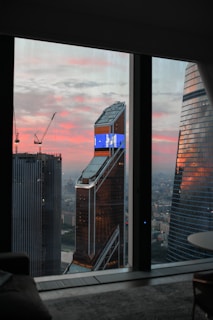 A view of a cityscape through a window, featuring tall modern skyscrapers with one building displaying a digital screen. The sky is painted with hues of pink and orange, indicating either sunrise or sunset. Construction cranes are visible, suggesting ongoing development.