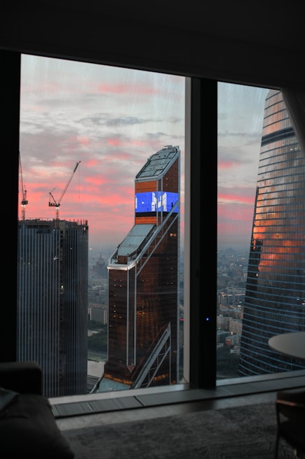 A view of a cityscape through a window, featuring tall modern skyscrapers with one building displaying a digital screen. The sky is painted with hues of pink and orange, indicating either sunrise or sunset. Construction cranes are visible, suggesting ongoing development.