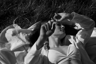 A person relaxes on grass, partly covered with a white blanket, wearing a light-colored tank top. Their eyes are closed, with an arm resting over their forehead, creating a peaceful and serene atmosphere.
