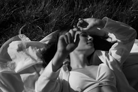 A person relaxes on grass, partly covered with a white blanket, wearing a light-colored tank top. Their eyes are closed, with an arm resting over their forehead, creating a peaceful and serene atmosphere.