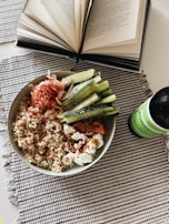 An open book with visible text lies on a table next to a bowl filled with quinoa, sliced cucumbers, and various toppings, including what appears to be smoked salmon and cream cheese. A green bottle of kombucha is placed beside the bowl. The setting is on a checkered fabric placemat.