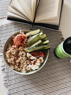 An open book with visible text lies on a table next to a bowl filled with quinoa, sliced cucumbers, and various toppings, including what appears to be smoked salmon and cream cheese. A green bottle of kombucha is placed beside the bowl. The setting is on a checkered fabric placemat.