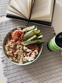 An open book with visible text lies on a table next to a bowl filled with quinoa, sliced cucumbers, and various toppings, including what appears to be smoked salmon and cream cheese. A green bottle of kombucha is placed beside the bowl. The setting is on a checkered fabric placemat.