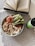 An open book with visible text lies on a table next to a bowl filled with quinoa, sliced cucumbers, and various toppings, including what appears to be smoked salmon and cream cheese. A green bottle of kombucha is placed beside the bowl. The setting is on a checkered fabric placemat.