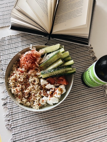 An open book with visible text lies on a table next to a bowl filled with quinoa, sliced cucumbers, and various toppings, including what appears to be smoked salmon and cream cheese. A green bottle of kombucha is placed beside the bowl. The setting is on a checkered fabric placemat.