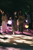 A group of people walking at night, each holding a glowing lantern that casts colorful lights and shadows on the ground. The atmosphere suggests a night event or festival, with attendees dressed in casual attire and some checking their phones. Trees and other attendees are visible in the dimly lit background.