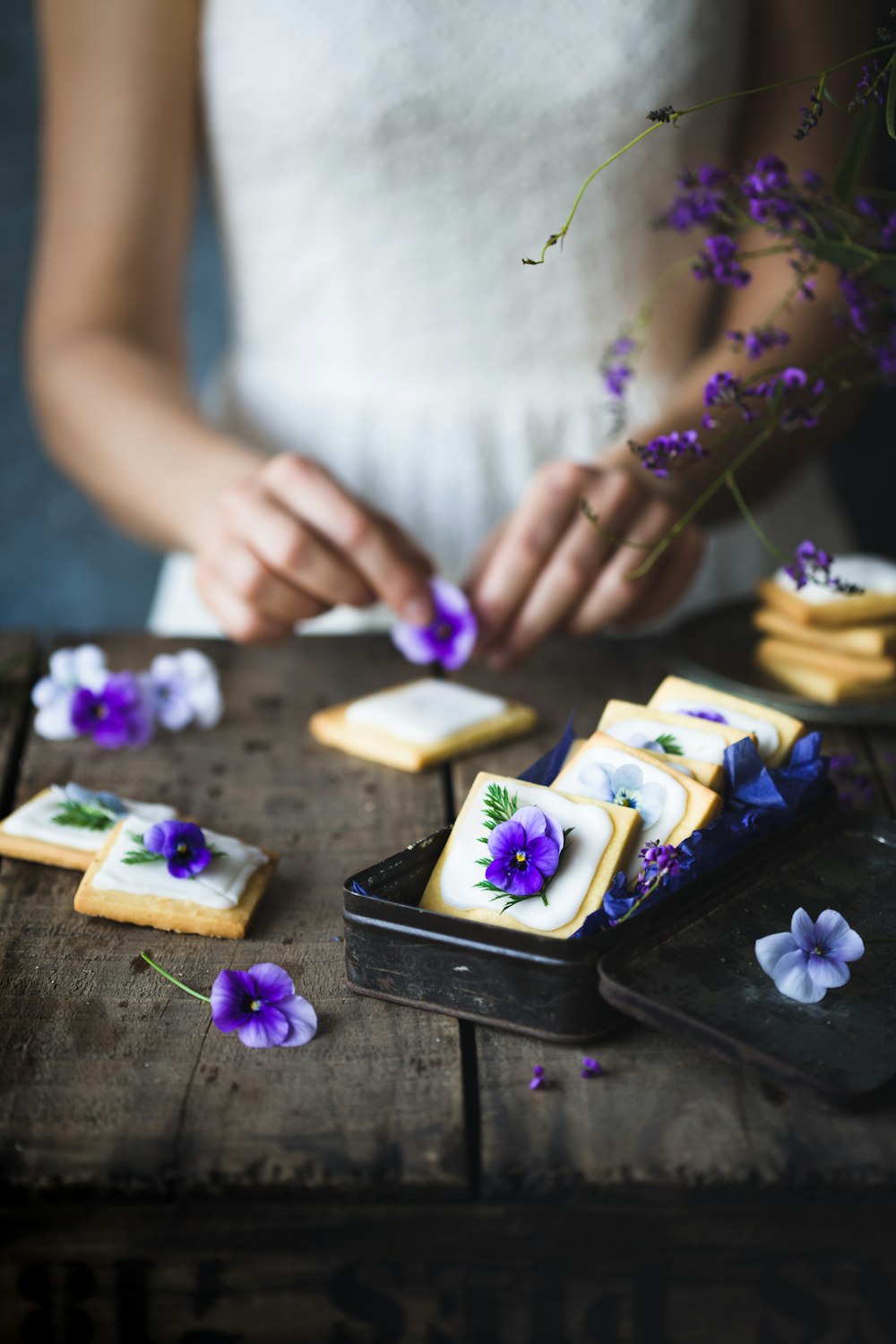 Une femme en robe blanche décore des biscuits avec des fleurs violettes