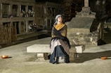A person wearing a shawl and headscarf sits on a stone bench in a dimly lit cemetery. The surroundings include tombstones, vases with flowers, and ornate metal fencing, suggesting a solemn and historical atmosphere.