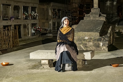 A person wearing a shawl and headscarf sits on a stone bench in a dimly lit cemetery. The surroundings include tombstones, vases with flowers, and ornate metal fencing, suggesting a solemn and historical atmosphere.