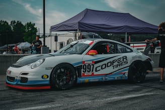 A white and blue race car with number 997 on its side is parked on a race track. It features prominent Cooper Tires branding. The car is surrounded by people and a tent in the background, indicating a racing event setting.