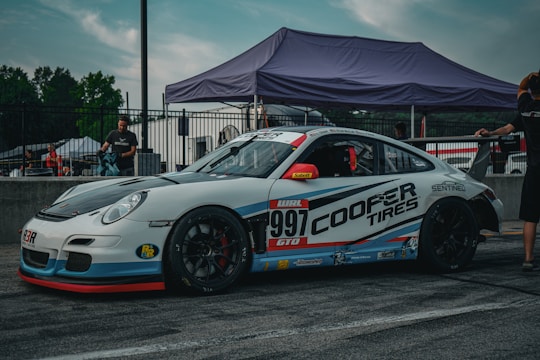 A white and blue race car with number 997 on its side is parked on a race track. It features prominent Cooper Tires branding. The car is surrounded by people and a tent in the background, indicating a racing event setting.