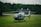 A blue and white helicopter is parked on a grassy field with a backdrop of dense green trees. Behind the helicopter, there is a rustic building with a dark roof and white chimneys. The sky is cloudy, creating a serene and peaceful atmosphere.