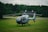 A blue and white helicopter is parked on a grassy field with a backdrop of dense green trees. Behind the helicopter, there is a rustic building with a dark roof and white chimneys. The sky is cloudy, creating a serene and peaceful atmosphere.