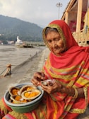 An elderly woman dressed in a vibrant red and yellow traditional Indian saree sits by a riverside. She is holding a matchbox and accompanied by a basket containing offerings of marigold flowers placed in leaf bowls. The background features a river, a distant statue, and misty hills.