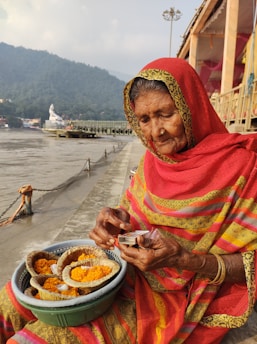 An elderly woman dressed in a vibrant red and yellow traditional Indian saree sits by a riverside. She is holding a matchbox and accompanied by a basket containing offerings of marigold flowers placed in leaf bowls. The background features a river, a distant statue, and misty hills.