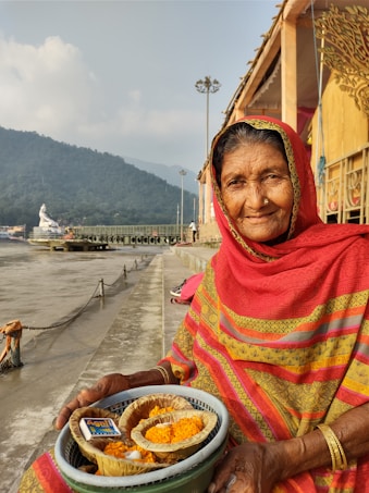 An elderly woman wearing a colorful sari is sitting by a riverbank, holding a basket containing marigold flowers and other items typically used for a ritual offering. In the background, hills and a cloudy sky are visible, along with a statue and a building structure on the riverbank.