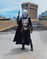 A person dressed in a detailed sci-fi costume stands on a tiled urban rooftop. The outfit includes a helmet with intricate designs, armor, a cape, and boots. A building and a city skyline are visible in the background under a clear sky.