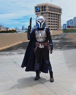 A person dressed in a detailed sci-fi costume stands on a tiled urban rooftop. The outfit includes a helmet with intricate designs, armor, a cape, and boots. A building and a city skyline are visible in the background under a clear sky.