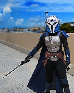 A person is dressed in a detailed, futuristic armor costume featuring a helmet with an owl-like design. The person is holding a sword with a unique pattern on the blade and is standing on a paved surface with a clear blue sky in the background.
