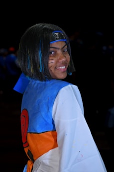 A person is wearing a costume inspired by an anime character with a blue and white garment, orange details, and an emblematic headband featuring a spiral symbol. The individual has face paint resembling whiskers and hair with blue streaks.