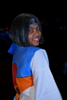 A person is wearing a costume inspired by an anime character with a blue and white garment, orange details, and an emblematic headband featuring a spiral symbol. The individual has face paint resembling whiskers and hair with blue streaks.