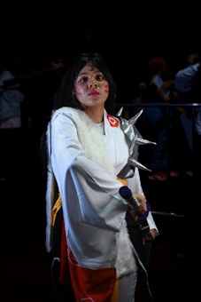 A person dressed in a white and red costume, possibly inspired by traditional or fantasy themes, stands confidently. They hold a prop resembling a sword and have facial makeup with red markings. The background is dark, with other figures visible in the shadows.