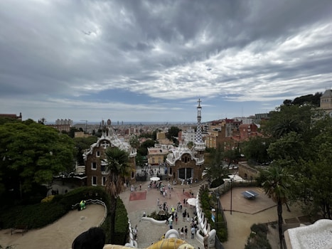 An expansive view of a cityscape with unique architectural structures in the foreground, possibly from Gaudí, situated in a verdant park setting. A cloudy sky looms above, and numerous people are gathered near the distinctive buildings.