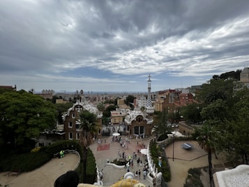 An expansive view of a cityscape with unique architectural structures in the foreground, possibly from Gaud&iacute;, situated in a verdant park setting. A cloudy sky looms above, and numerous people are gathered near the distinctive buildings.