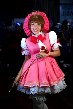A person is dressed in a vibrant pink and white costume with a large pink bonnet and a matching bow. The outfit includes intricate details like lace and gold trim. They are holding a wand with a red circular top and feather-like decorations. The background is dark, with blurred figures in the distance, suggesting a crowded event.