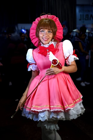A person is dressed in a vibrant pink and white costume with a large pink bonnet and a matching bow. The outfit includes intricate details like lace and gold trim. They are holding a wand with a red circular top and feather-like decorations. The background is dark, with blurred figures in the distance, suggesting a crowded event.