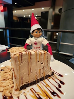 A toy figure dressed in a festive outfit with a red hat and a shirt that reads 'Snow' is positioned behind a large slice of cake topped with nuts and drizzled with chocolate and caramel sauce. The setting appears to be indoors with modern decor and ambient lighting.