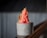 A creamy, pink sorbet is served in a simple, textured gray cup. The sorbet is garnished with a thin round slice of dried lime and small zest pieces, adding a touch of green color. The background features a blurred, corrugated texture.